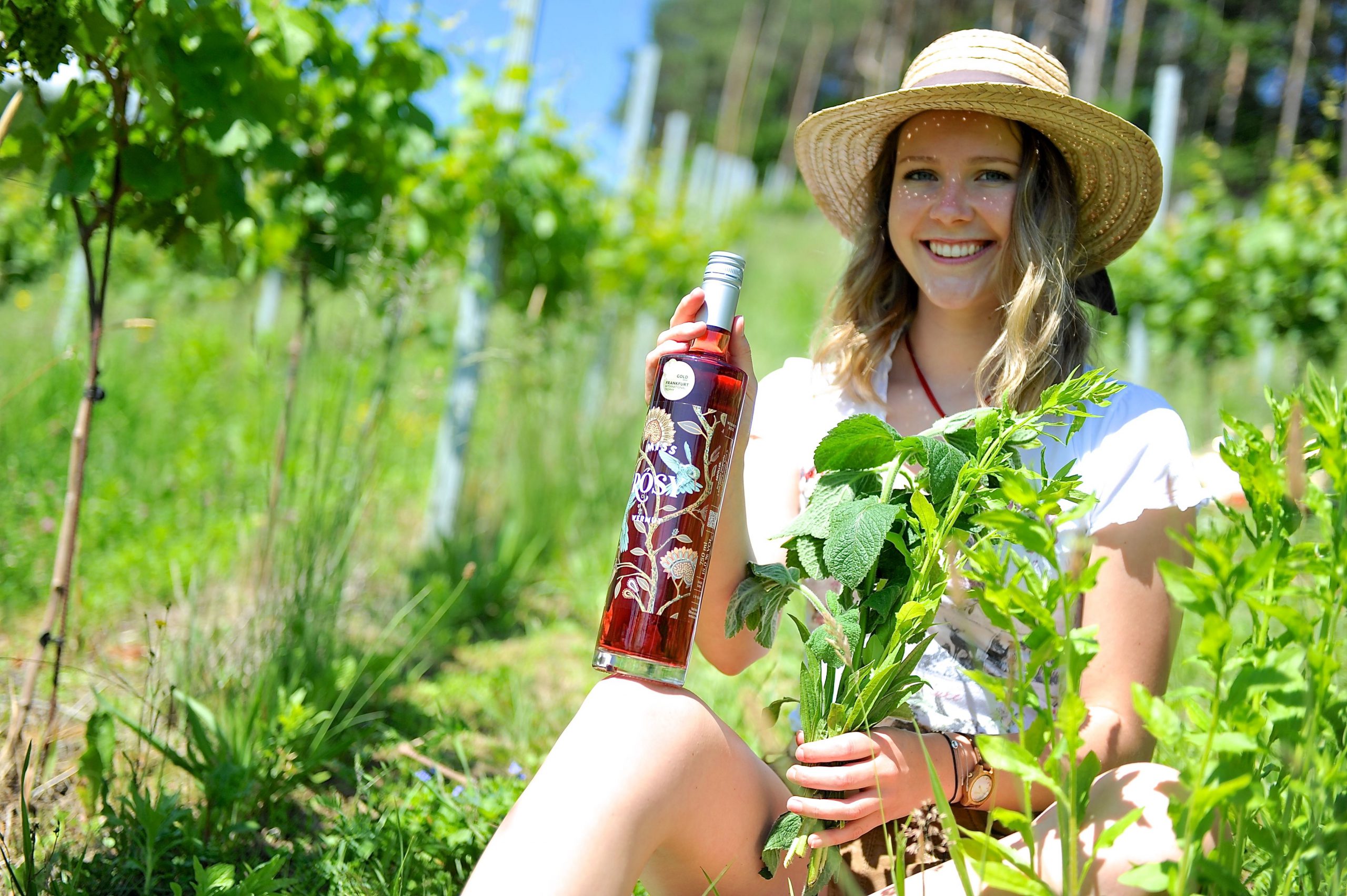 Junge Frau im Weingarten mit Kräutern und einer Flasche