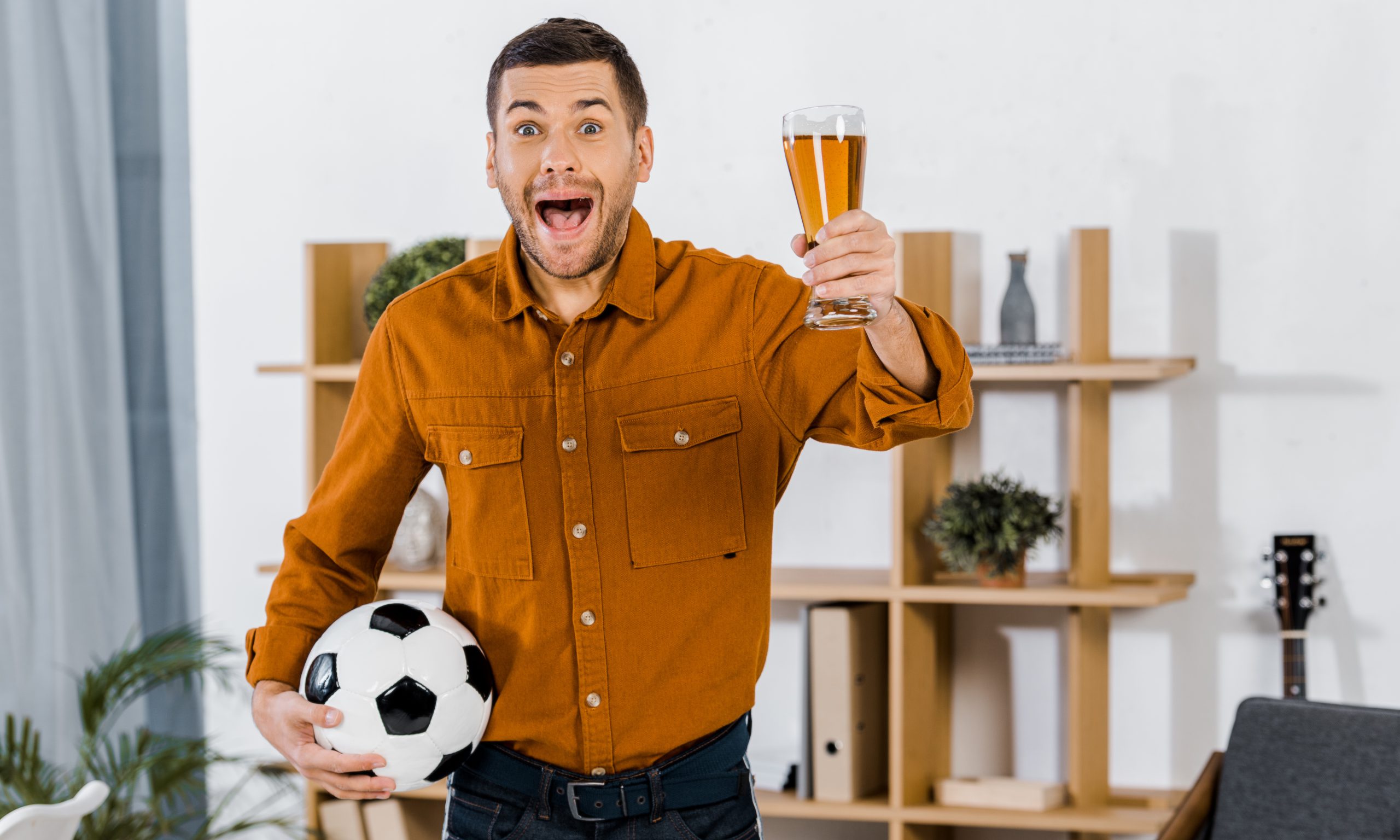 schöner Mann in modernem Wohnzimmer schreit, während er Fußball und Bier in der Hand hält